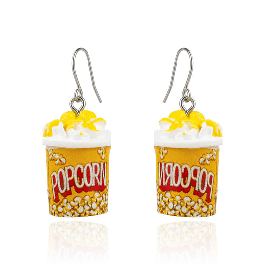 A pair of novelty popcorn bucket earrings with yellow and white popcorn, white whipped cream, and a titanium fishhook ear wire, displayed on a clean white background with a subtle reflection.