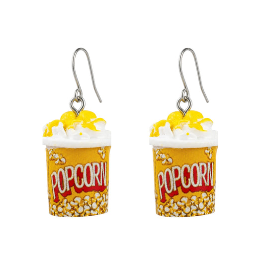 A pair of novelty popcorn bucket earrings with yellow and white popcorn, white whipped cream, and a titanium fishhook ear wire, displayed on a clean white background with a subtle reflection.