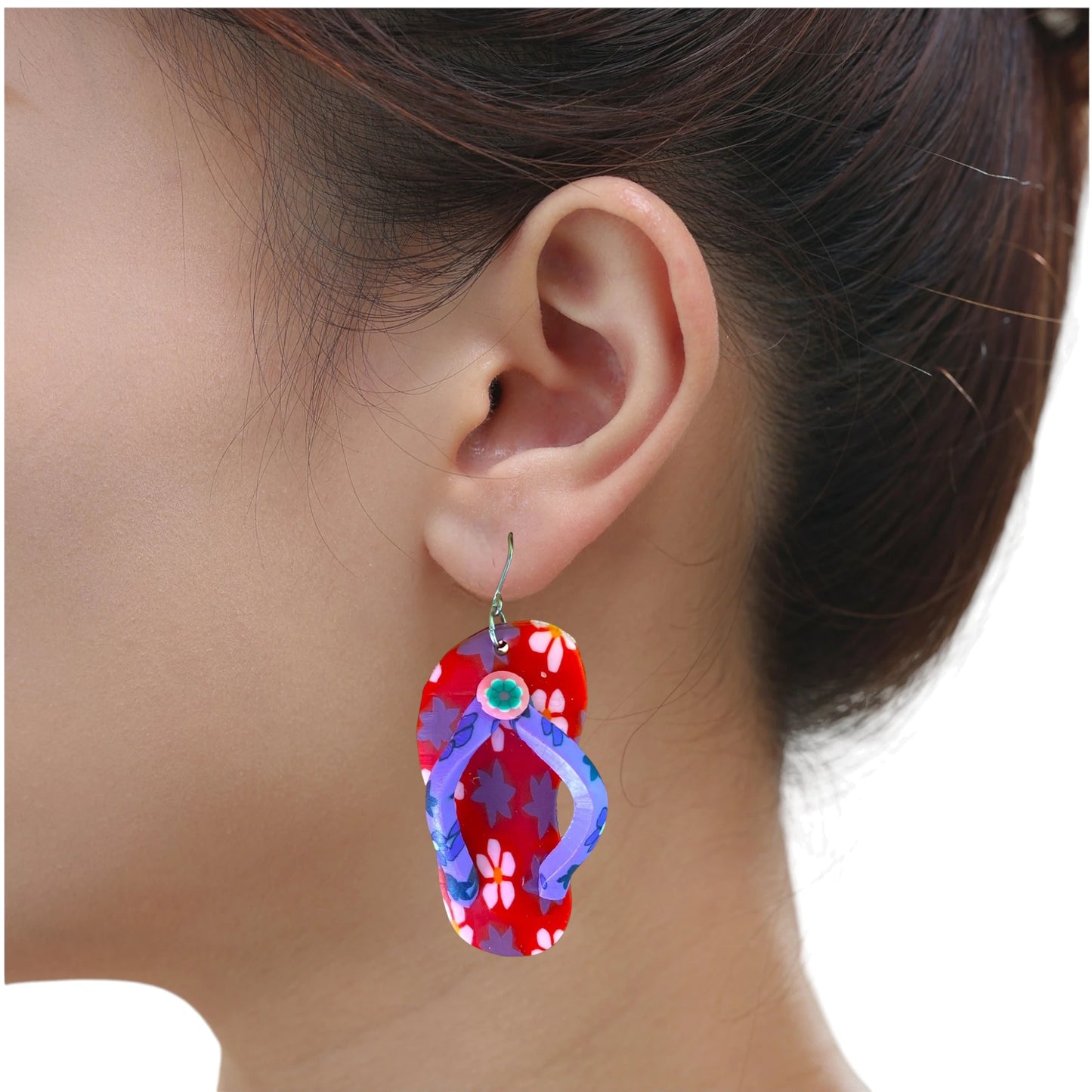 A close-up of a person's ear wearing a single titanium flip-flop-shaped dangling earring. The earring has a red floral upper with blue straps and a small green circular detail at the top, shown against a white background.