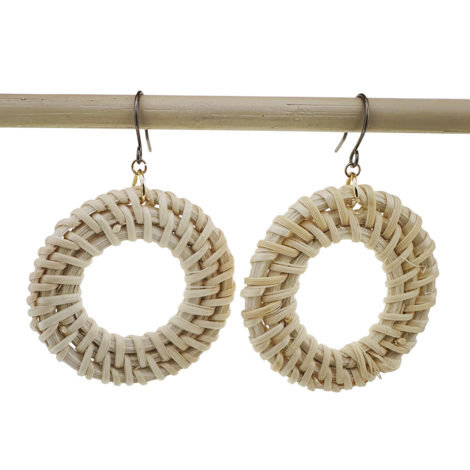 A pair of large circular woven straw earrings hanging from titanium fishhook wires, displayed against a clean white background with a light wood rod behind them.