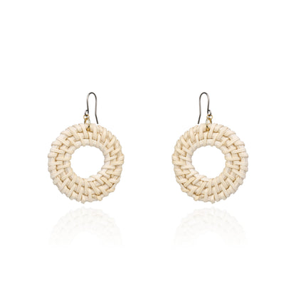 A pair of handmade circular woven earrings made from natural beige raffia or straw, suspended from titanium fishhook wires, shown on a clean white background with a soft reflection below.