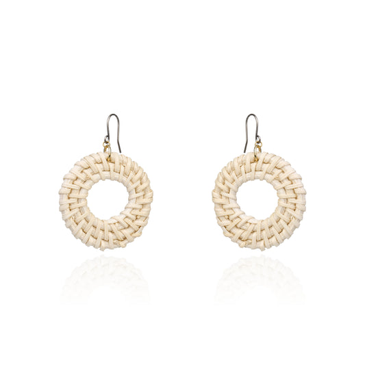 A pair of handmade circular woven earrings made from natural beige raffia or straw, suspended from titanium fishhook wires, shown on a clean white background with a soft reflection below.