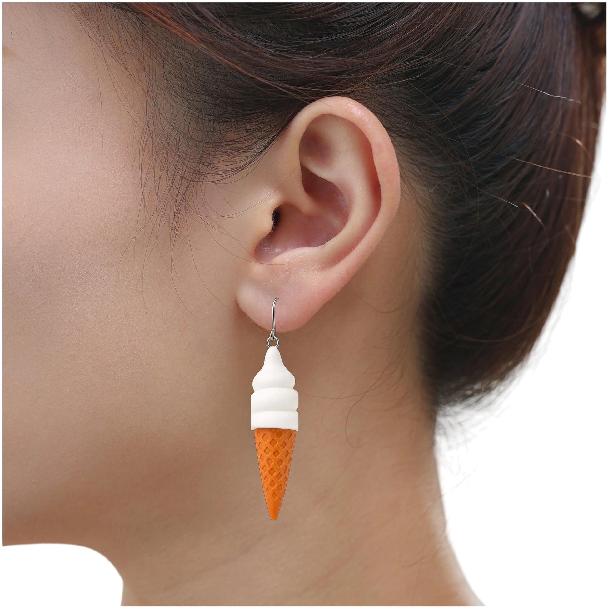 A close-up of a person's ear wearing a dangling ice cream cone earring. The earring has a white soft-serve top and an orange waffle-cone bottom on a titanium hook.