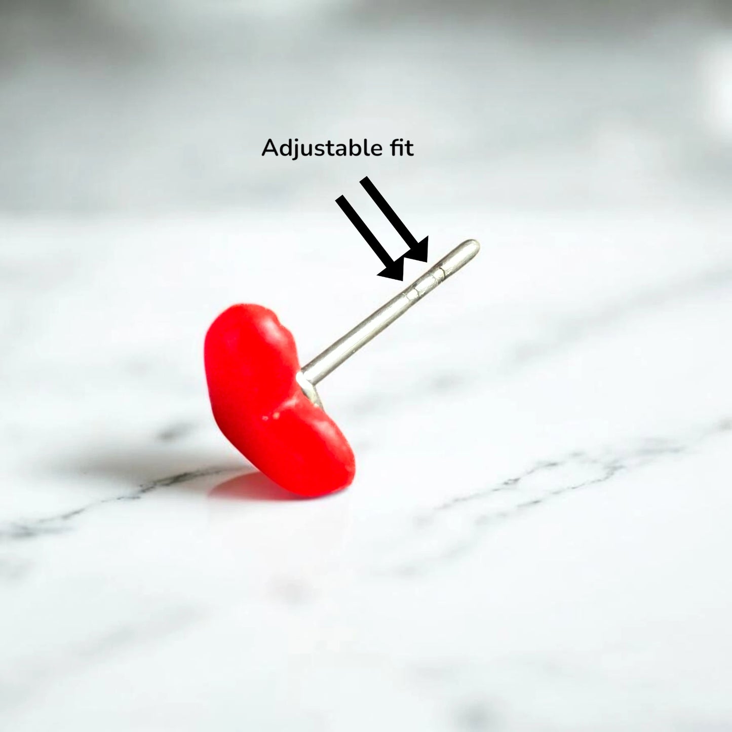 A red curved stud earring with a silver adjustable titanium post, shown on a white marble surface with an 'Adjustable fit' text overlay and arrows pointing to the sizing rings.