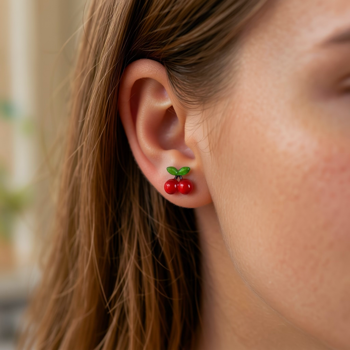 A girl wearing cherry-themed stud earrings with red and green details and titanium post and backs, shown on a  beige background.