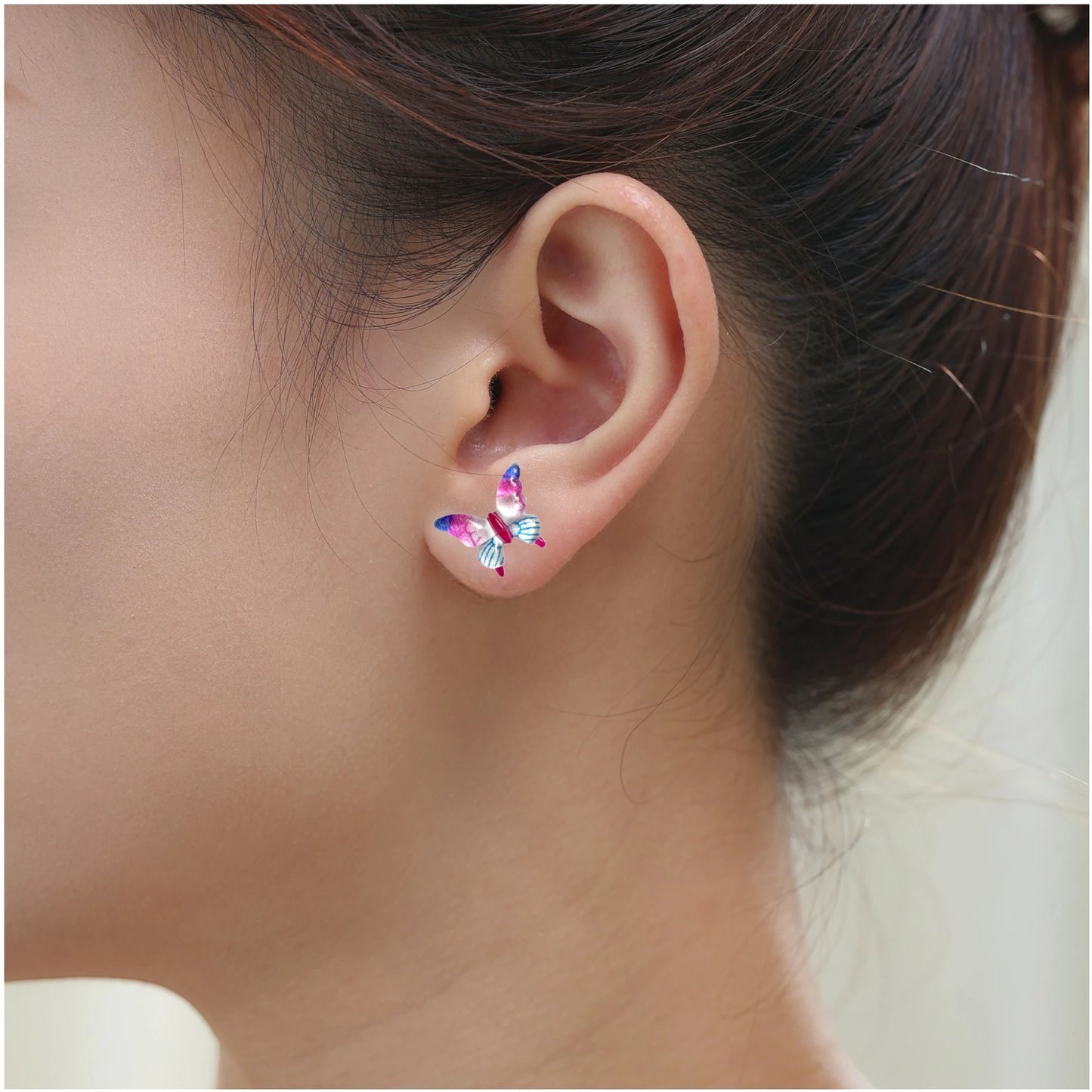 A close-up of a person's ear wearing a small butterfly-shaped titanium stud earring in shades of pink, blue.