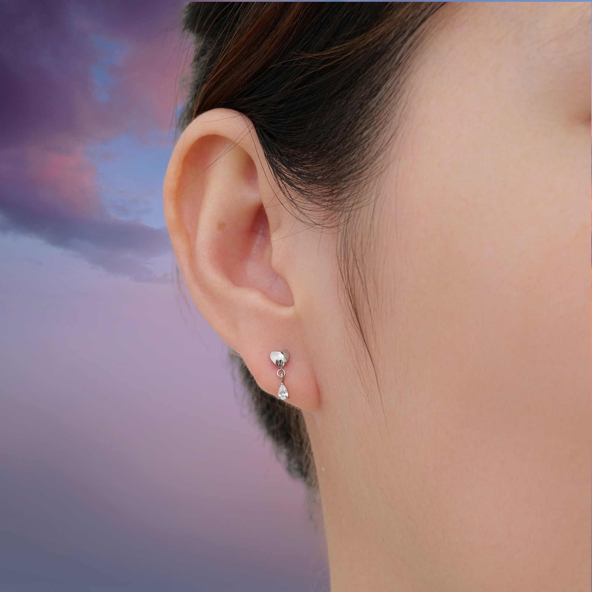 Titanium earring with heart and teardrop hanging gem worn by a woman with a sky background