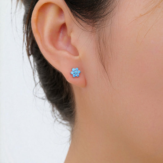Close-up of an ear wearing a titanium threadless slat back floral aqua blue stud earring with a blurred background