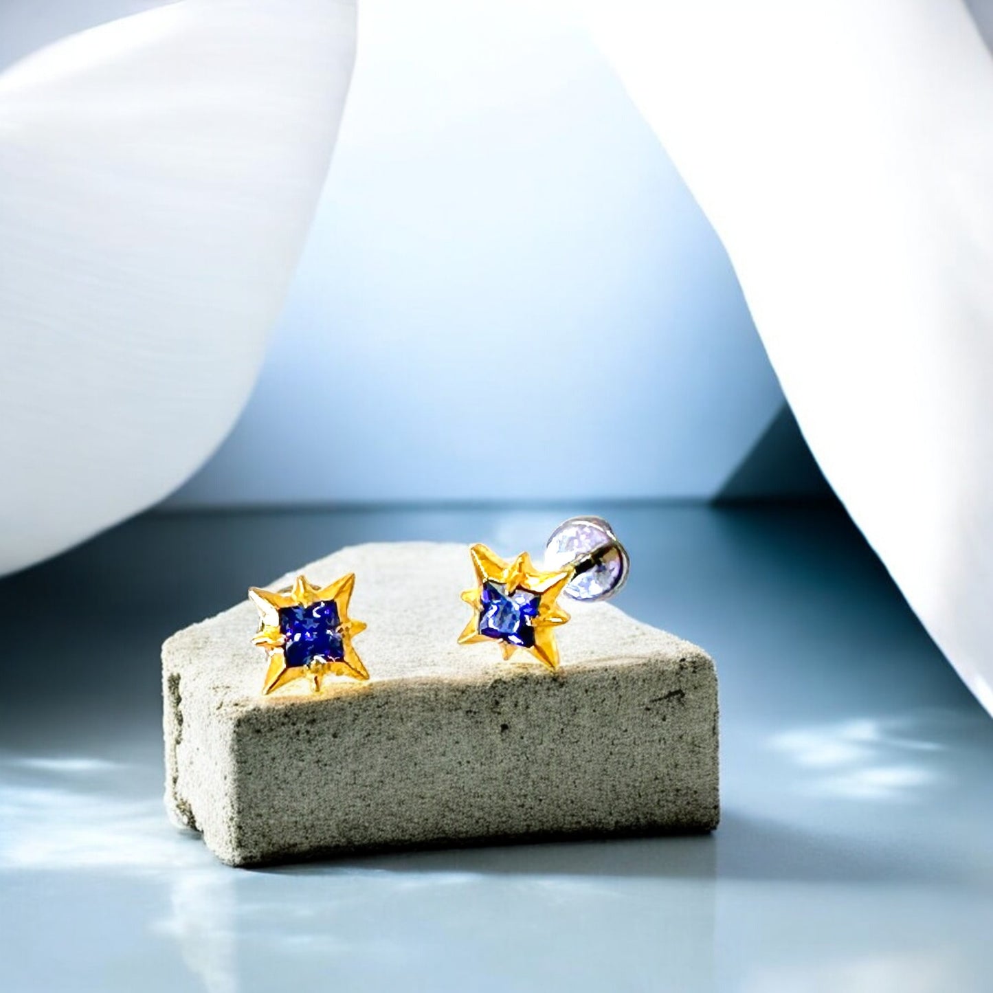 Star-shaped titanium flat back earrings with blue gemstones and gold accents on a stone surface.